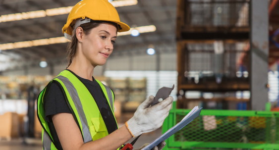 A person wearing a yellow hard hat, safety vest, and gloves inspects an item while holding a clipboard inside an industrial warehouse