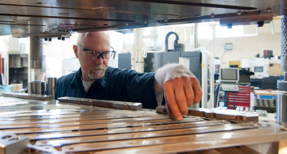 A person wearing safety glasses inspects and adjusts metal components inside a tool and die or moldmaking machine in a manufacturing workshop
