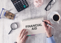 man's hand writing financial aid on notebook with calculator, money and other desk accessories