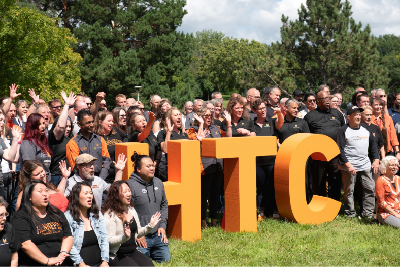 Hennepin Tech employees gathered outdoors around large, orange "HTC" letters, cheering.