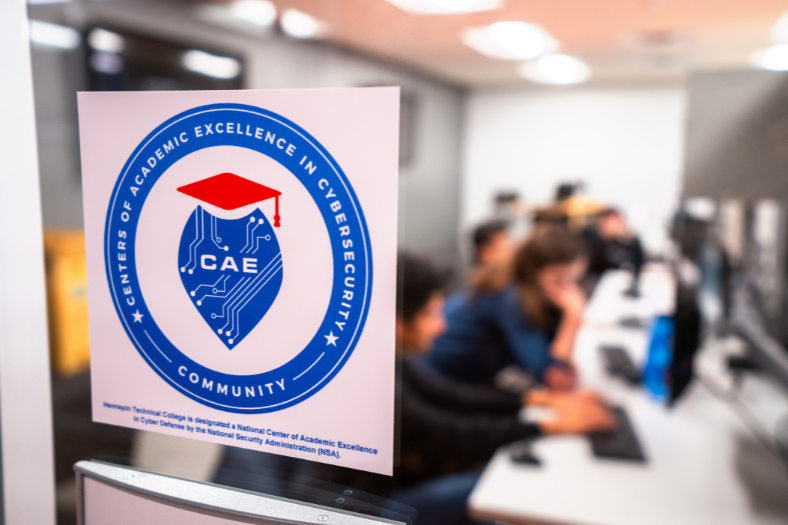 A blue and red circular crest reading "Center of Academic Excellence in Cybersecurity" is featured prominently on the left of the image. In the background, a group of blurred students work on computers